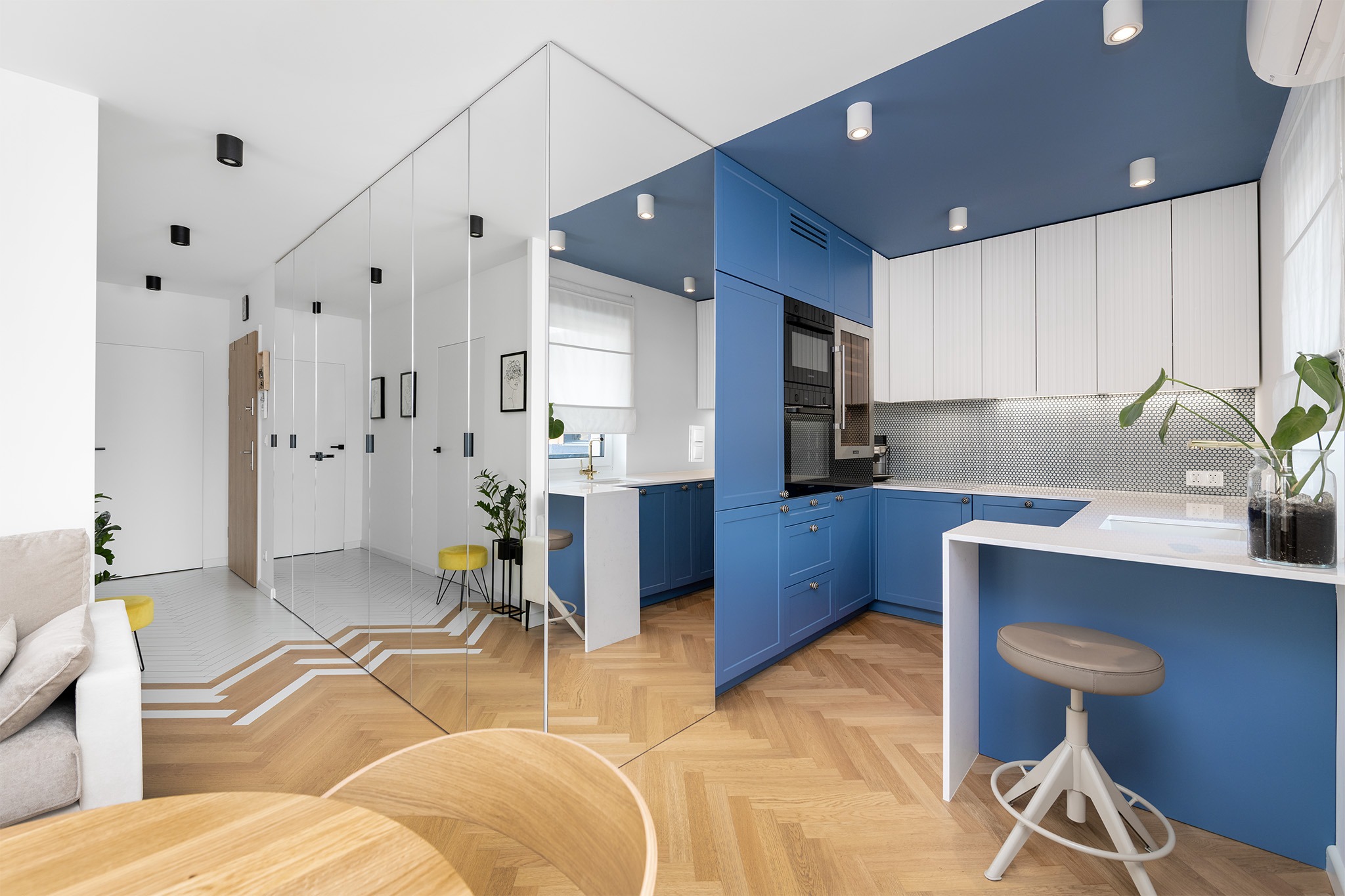 Nowoczesna kuchnia z niebieskimi szafkami i białymi górnymi szafkami, odbijająca się w lustrzanej ścianie, ukazująca przestronne wnętrze mieszkania.