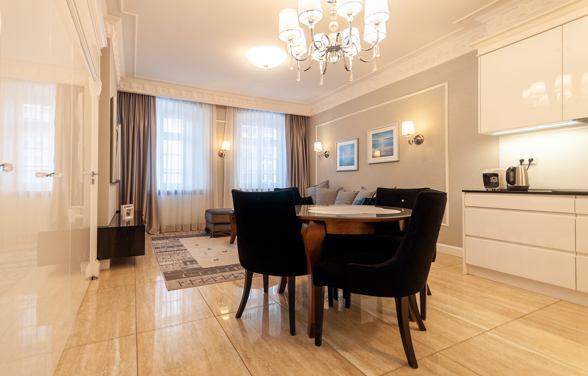 Eleganckie wnętrze mieszkania: połyskująca podłoga z płytek, okrągły drewniany stół z czarnymi krzesłami, w tle sofa z poduszkami i kryształowy żyrandol pod sufitem z dekoracyjną sztukaterią.