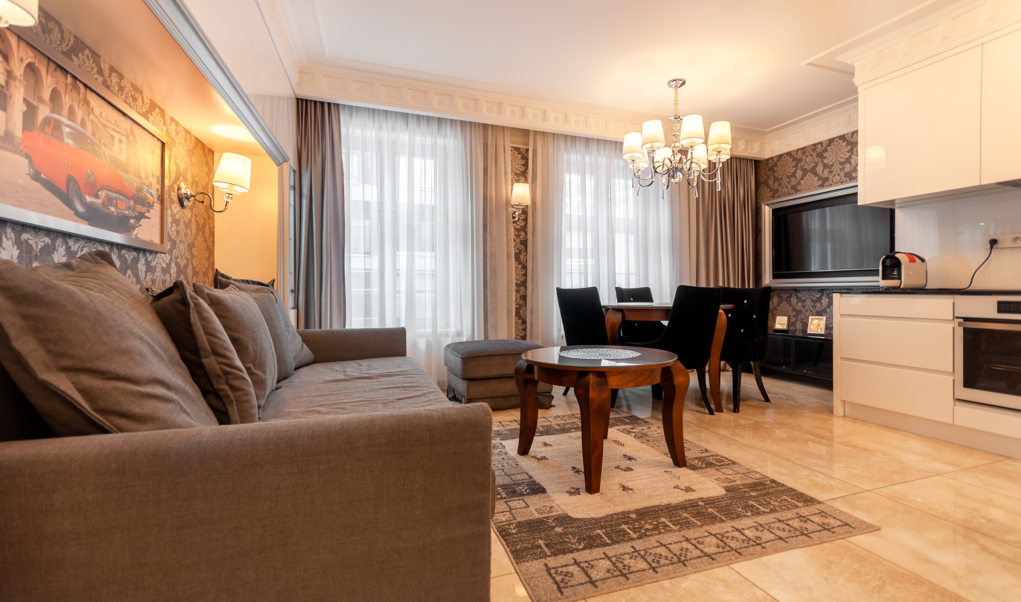 Salon z aneksem kuchennym w stylu klasycznym: szary narożnik z poduszkami, okrągły stolik kawowy na dywanie, stół z krzesłami, biała zabudowa kuchenna, telewizor na ścianie z wzorzystą tapetą...