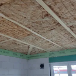 Poddasze w trakcie prac izolacyjnych, widoczna wełna mineralna między drewnianymi belkami, zielone elementy Lohrelement na ścianach z betonu komórkowego, okno w tle.