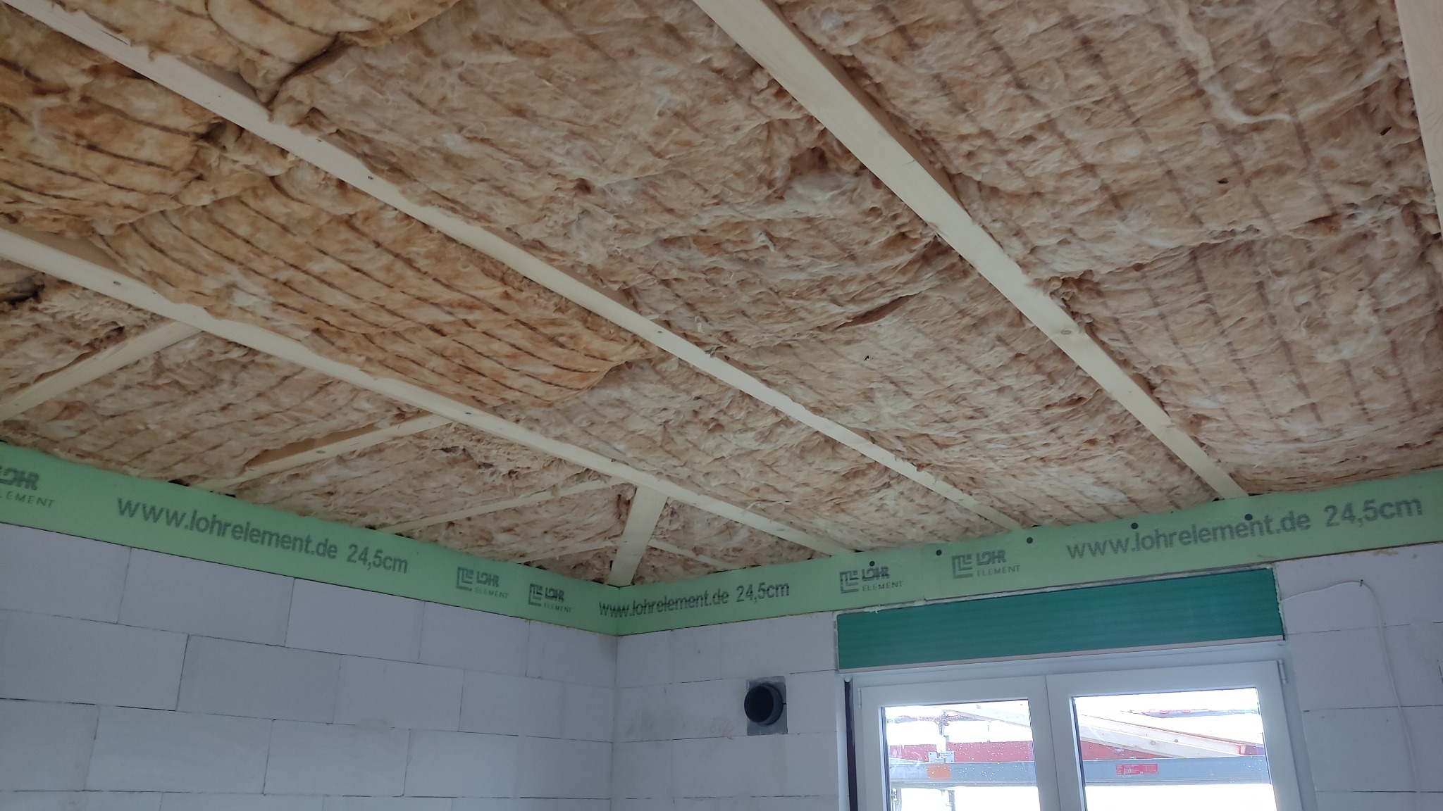 Poddasze w trakcie prac izolacyjnych, widoczna wełna mineralna między drewnianymi belkami, zielone elementy Lohrelement na ścianach z betonu komórkowego, okno w tle.