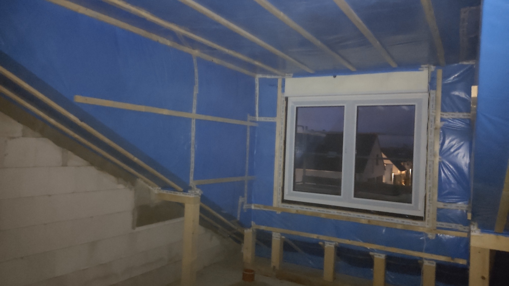 Poddasze w trakcie ocieplania: widoczne drewniane elementy konstrukcyjne, folia paroizolacyjna w kolorze niebieskim, okno z widokiem na domy w tle, ściana z białych bloczków.