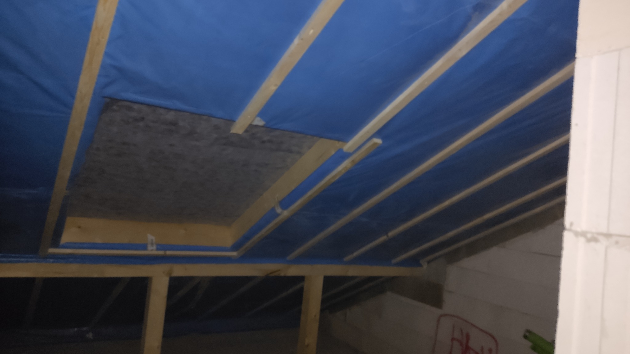 Poddasze w trakcie izolacji: widoczne drewniane krokwie, niebieska membrana paroizolacyjna oraz fragment niezabezpieczonej przestrzeni, prawdopodobnie otworu rewizyjnego.