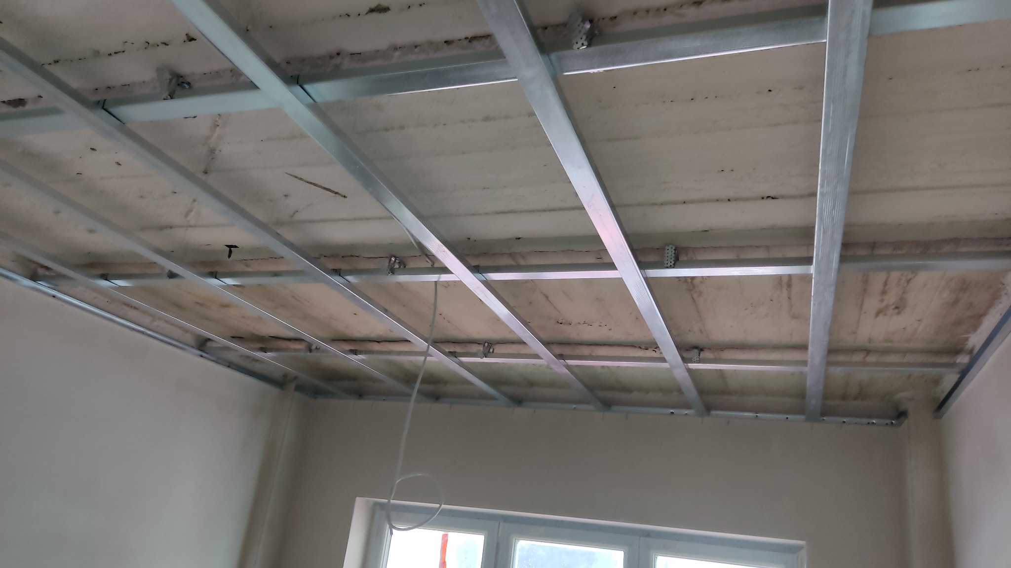 Stelaż sufitowy z metalowych profili zamocowany do surowego stropu, przygotowany do montażu płyt, widok z dołu, fragment okna w tle.