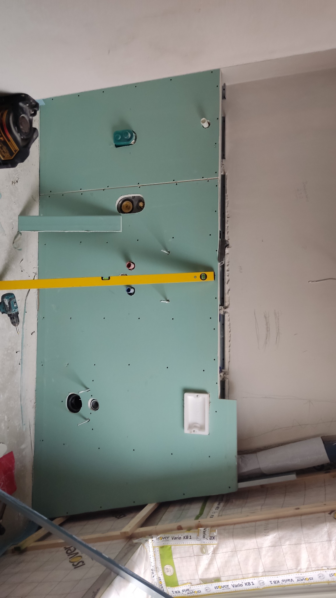 Montaż płyty karton gips w łazience, widoczne otwory na instalacje wodno-kanalizacyjne, poziomica i wkrętarka, izolacja podłogi.
