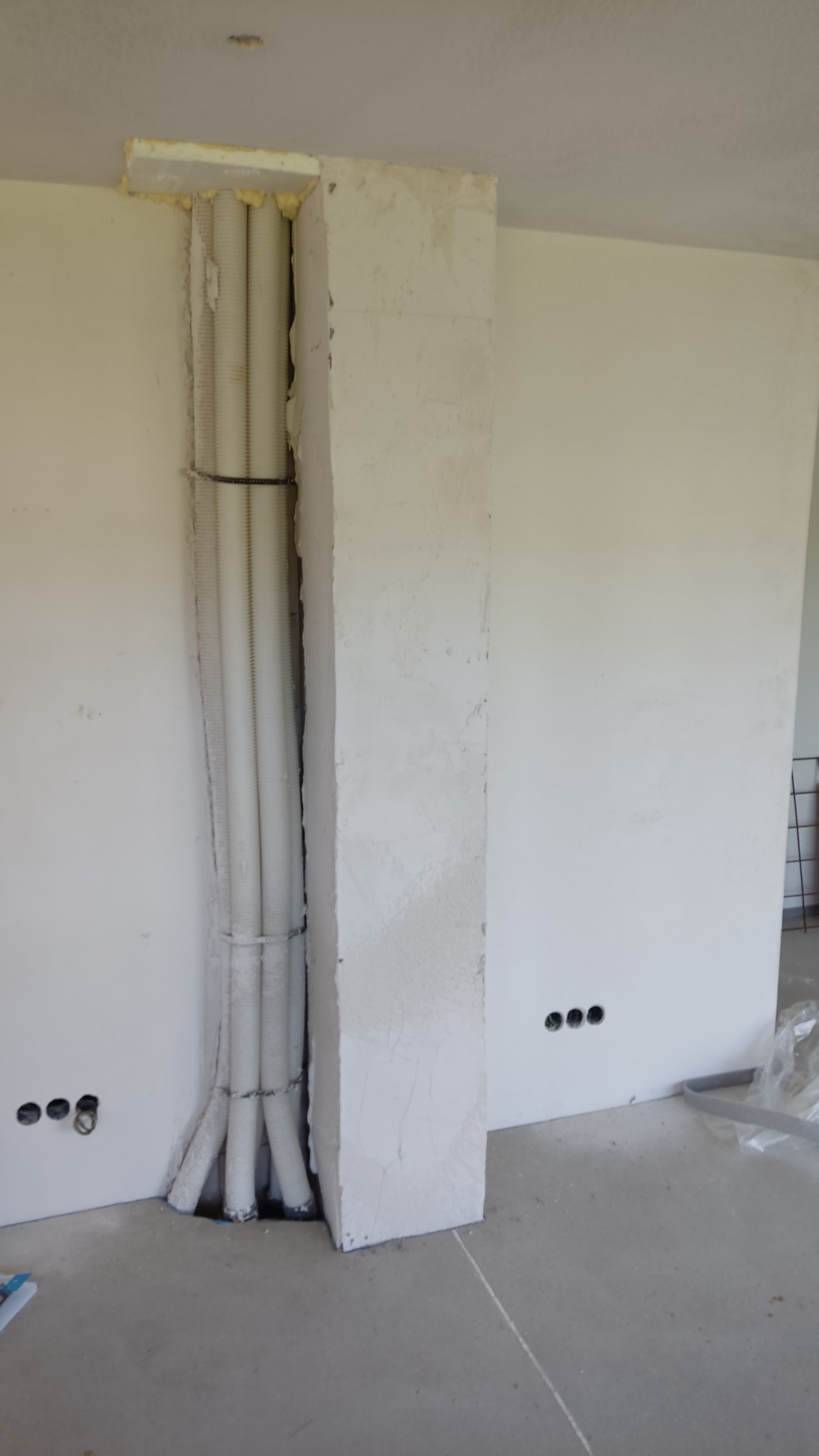 Wnęka w ścianie z pionowo ułożonymi rurami w karbowanej osłonie, zabezpieczonymi opaskami, z widocznymi śladami po szpachlowaniu i przygotowaniu do dalszych prac wykończeniowych.