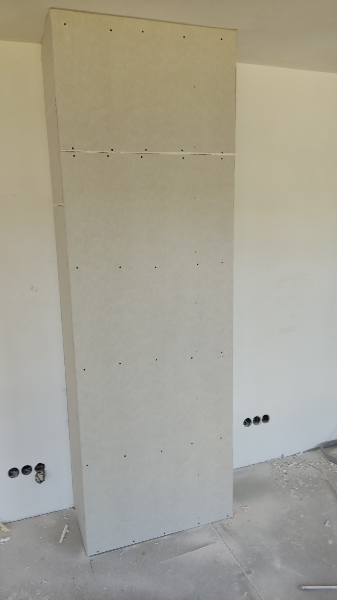 Wzniesiona konstrukcja z płyt na ścianie, widoczne ślady po wkrętach, przygotowana do dalszych prac wykończeniowych, z widocznymi otworami na instalacje elektryczne w ścianie obok.