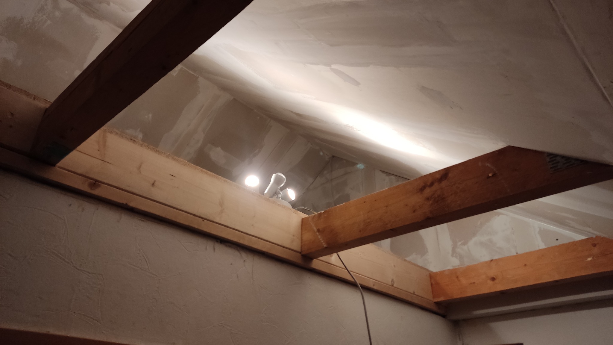 Poddasze w trakcie prac wykończeniowych, widoczne drewniane belki konstrukcyjne, oświetlenie robocze i ślady świeżo nałożonej masy szpachlowej na ścianach i suficie.