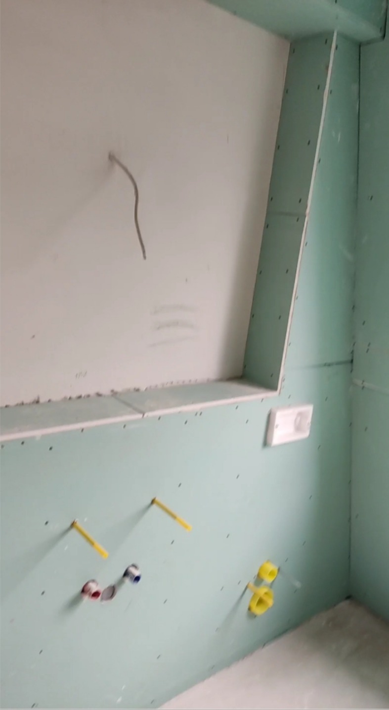 Ściana w trakcie zabudowy, widoczne płyty gipsowo-kartonowe, wystające rury instalacji wodnej z zaślepkami, kabel elektryczny i puszka instalacyjna, narożnik wykończony płytą.