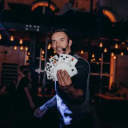 Uśmiechnięty iluzjonista z mikrofonem prezentuje wachlarz kart na tle industrialnego wnętrza z żarówkami.