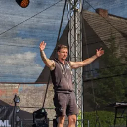 Mężczyzna w czarnej bluzie z kapturem i tatuażem na ramieniu stoi na scenie z podniesionymi rękami, za nim konstrukcja sceniczna z oświetleniem i czarną siatką.