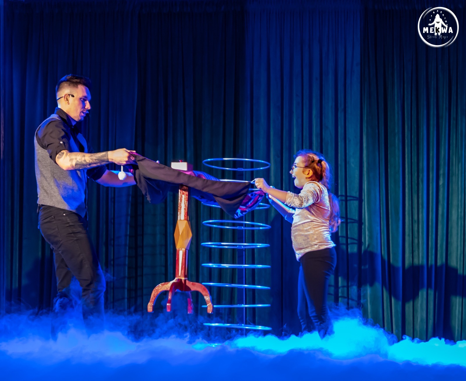 Iluzjonista z tatuażami, asystuje mu uśmiechnięta dziewczynka podczas lewitacji stolika na tle niebieskich kurtyn i dymu scenicznego.
