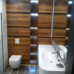 Nowoczesna łazienka z zabudową z drewnopodobnej płyty, czarnymi akcentami, podświetlanymi półkami, białą umywalką i toaletą oraz marmurową podłogą.