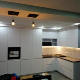 Nowoczesna, minimalistyczna kuchnia z białymi frontami, czarnym piekarnikiem i płytą gazową, oświetlona wiszącymi żarówkami edisonowskimi i ledowym pasem pod szafkami górnymi, z fragmentem salonu...