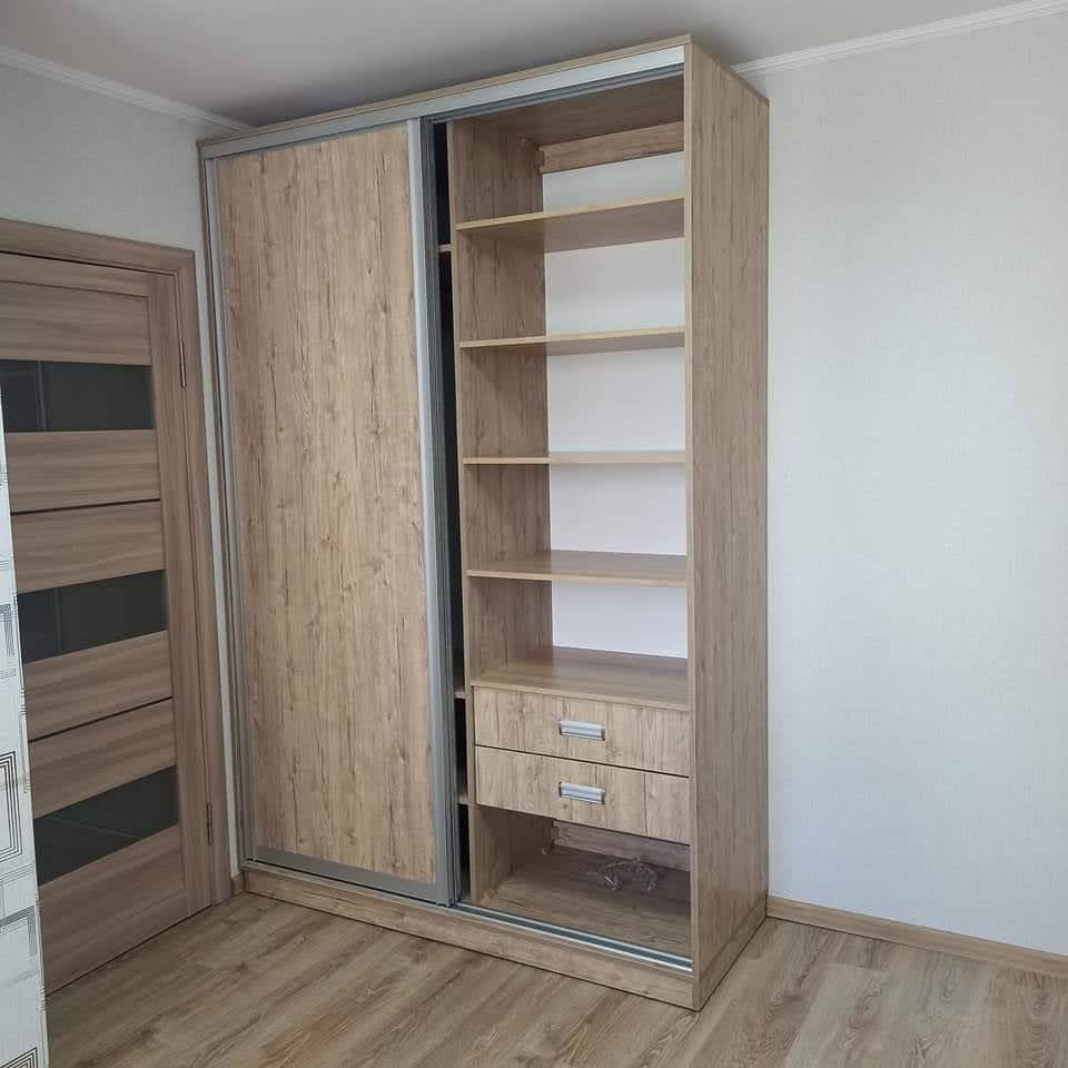 Beżowa szafa przesuwna z drzwiami z imitacją drewna i otwartymi półkami z szufladami, widoczna częściowo odsłonięta zawartość półek, na podłodze panele podłogowe w odcieniach brązu.