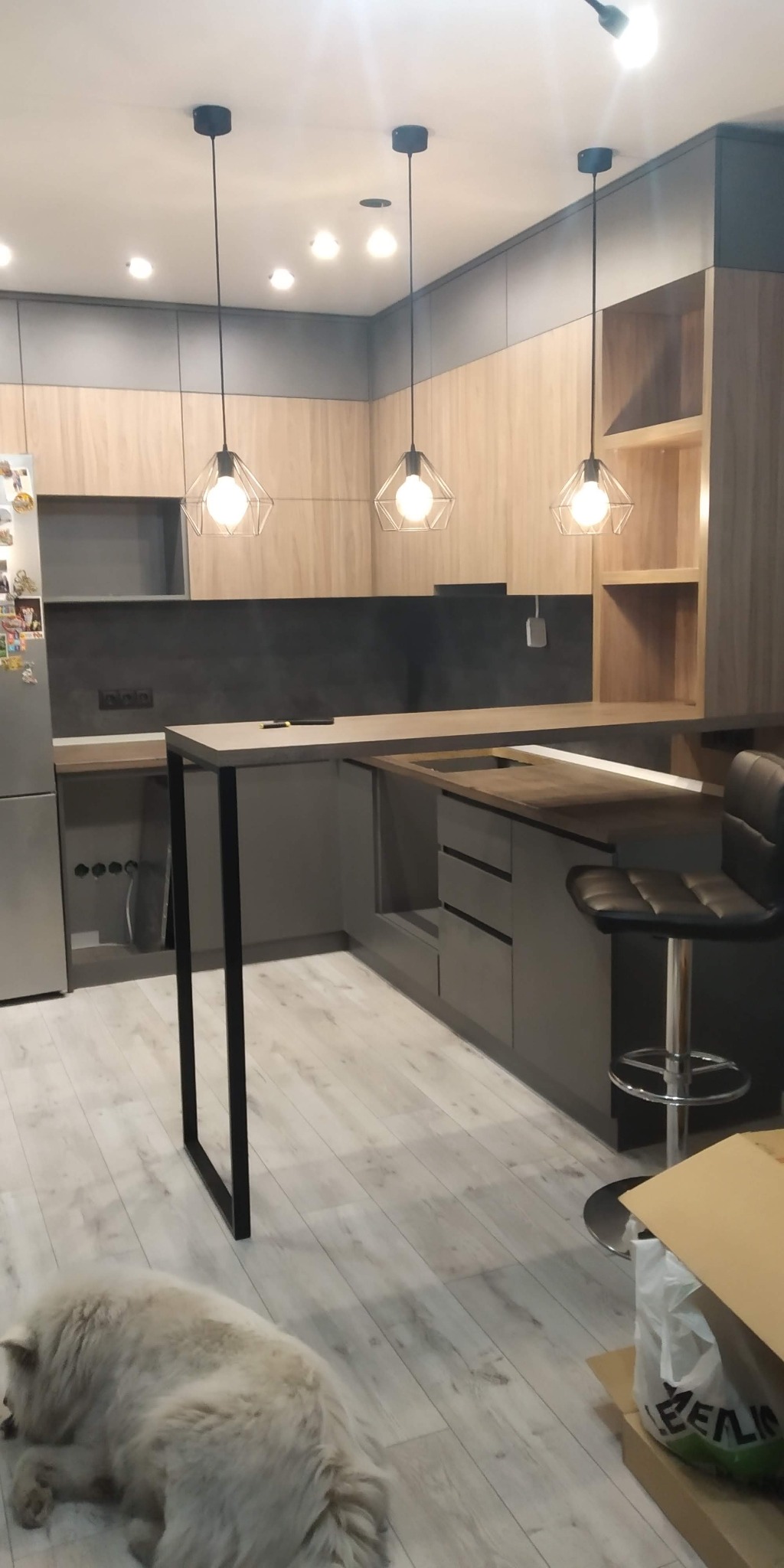 Nowoczesna kuchnia z drewnianymi frontami szafek i szarymi elementami, wyspą z czarnymi nogami i trzema lampami wiszącymi o geometrycznych kloszach. Na podłodze leży biały pies.