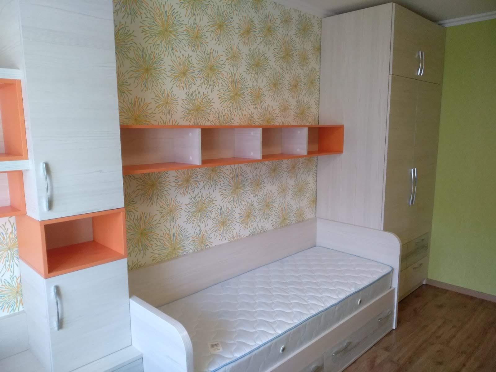 Pokój młodzieżowy z zabudową meblową: szafa, półki wiszące w kolorze pomarańczowym, szafki wiszące i łóżko z szufladami, wszystko w jasnym drewnie z widoczną strukturą, na ścianie tapeta w kwiaty.