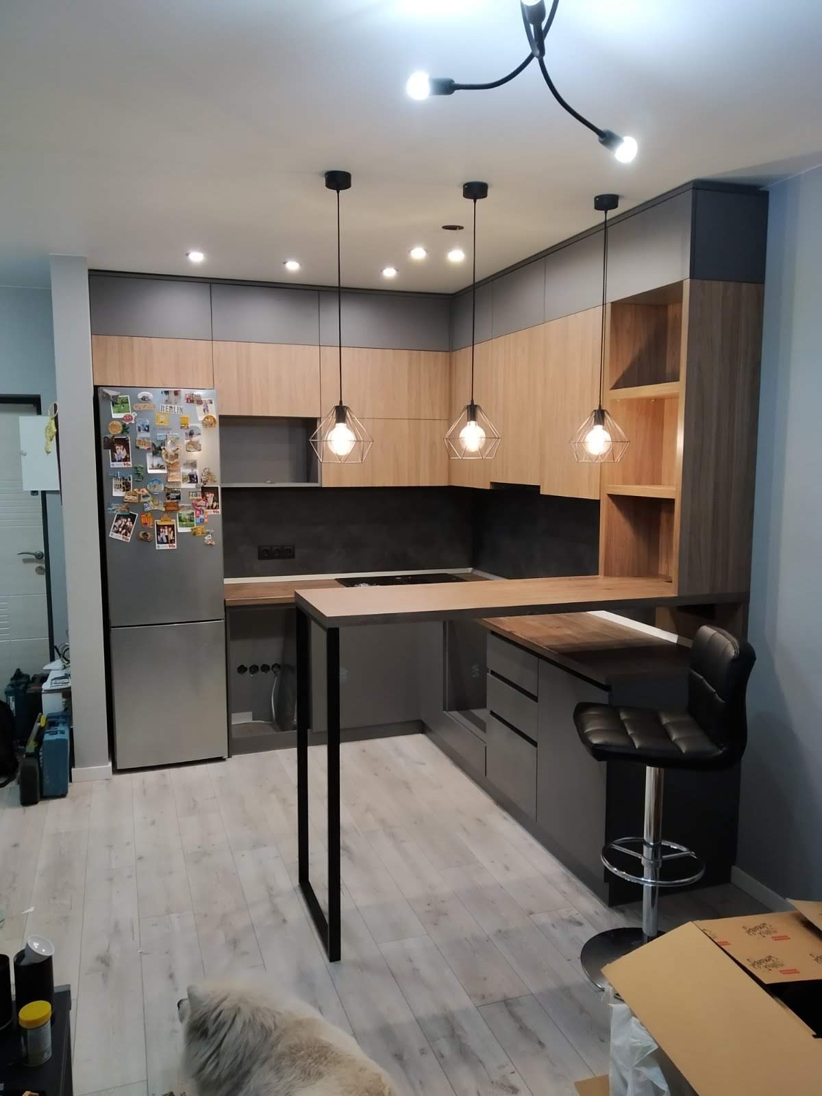 Nowoczesna kuchnia z szarymi i drewnianymi frontami, wyspą z czarną ramą, oświetleniem wiszącym w stylu industrialnym i lodówką obklejoną magnesami, widoczny pies leżący na podłodze.