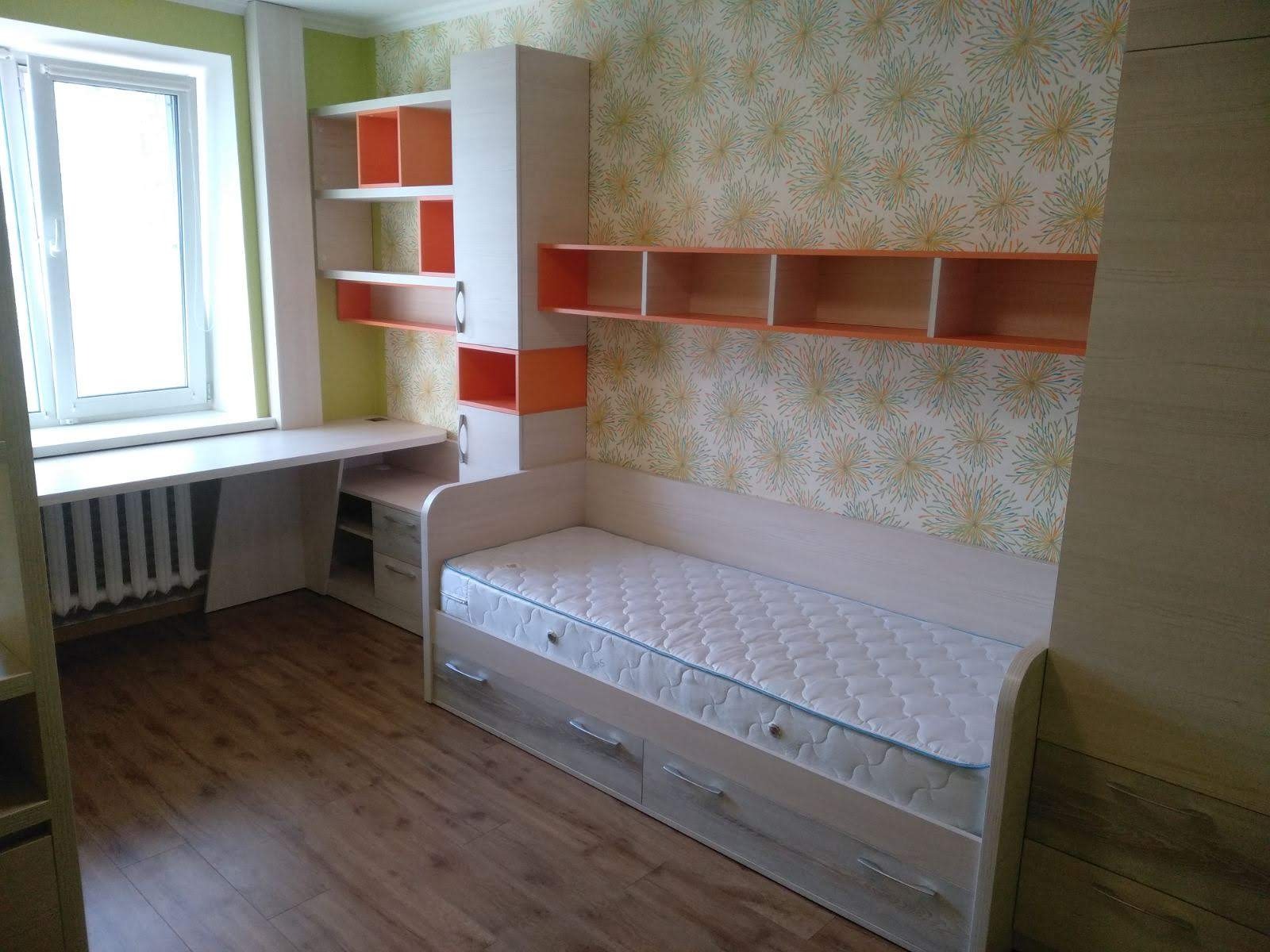 Pokój dziecięcy z łóżkiem z szufladami, biurkiem pod oknem i regałem w kolorze kremowym z pomarańczowymi akcentami na tle tapety w kwiaty.