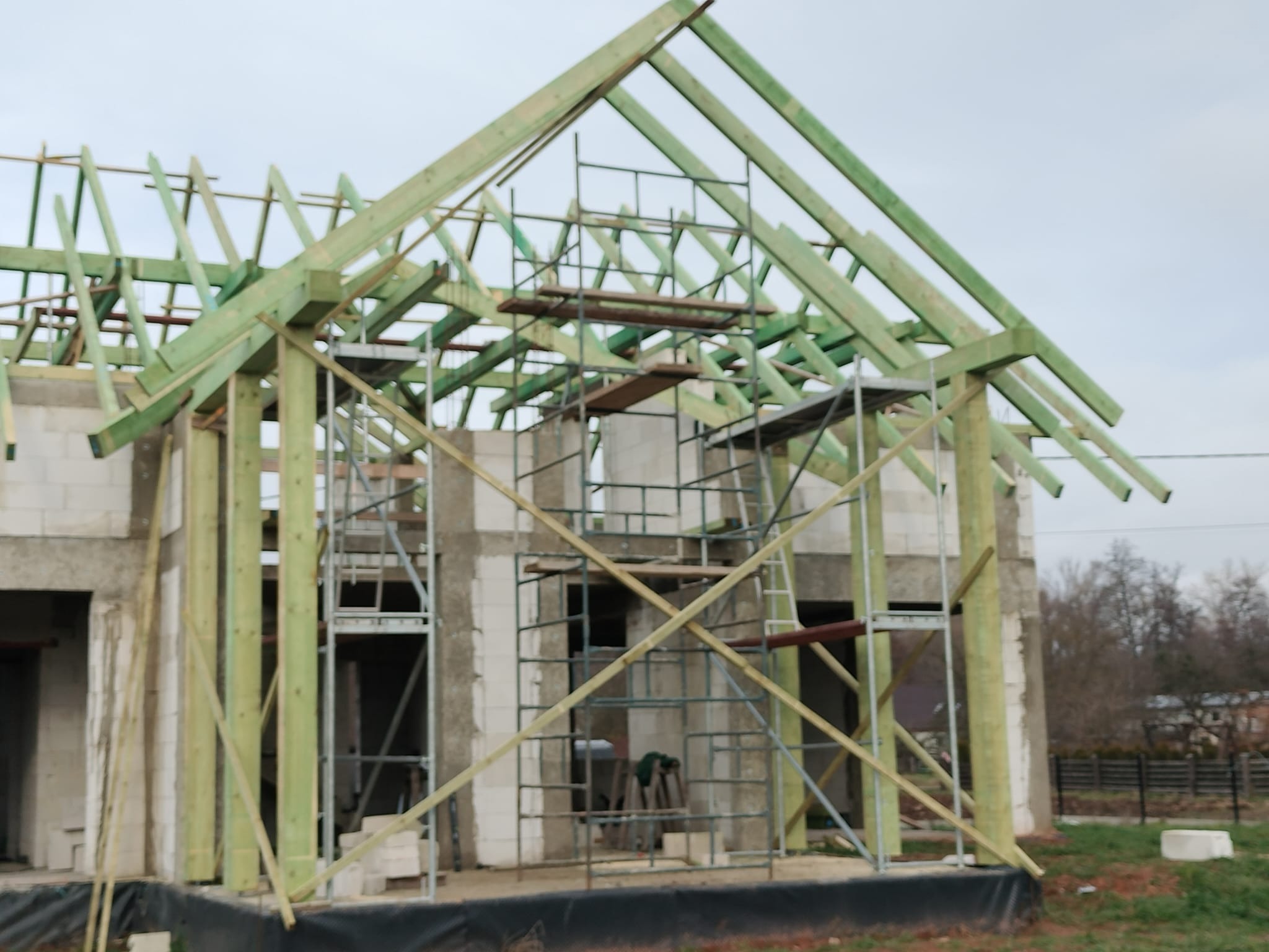 Wznoszenie konstrukcji dachu z zielonych belek na surowym budynku z białych bloczków. Widoczne rusztowania i elementy więźby dachowej w trakcie montażu.
