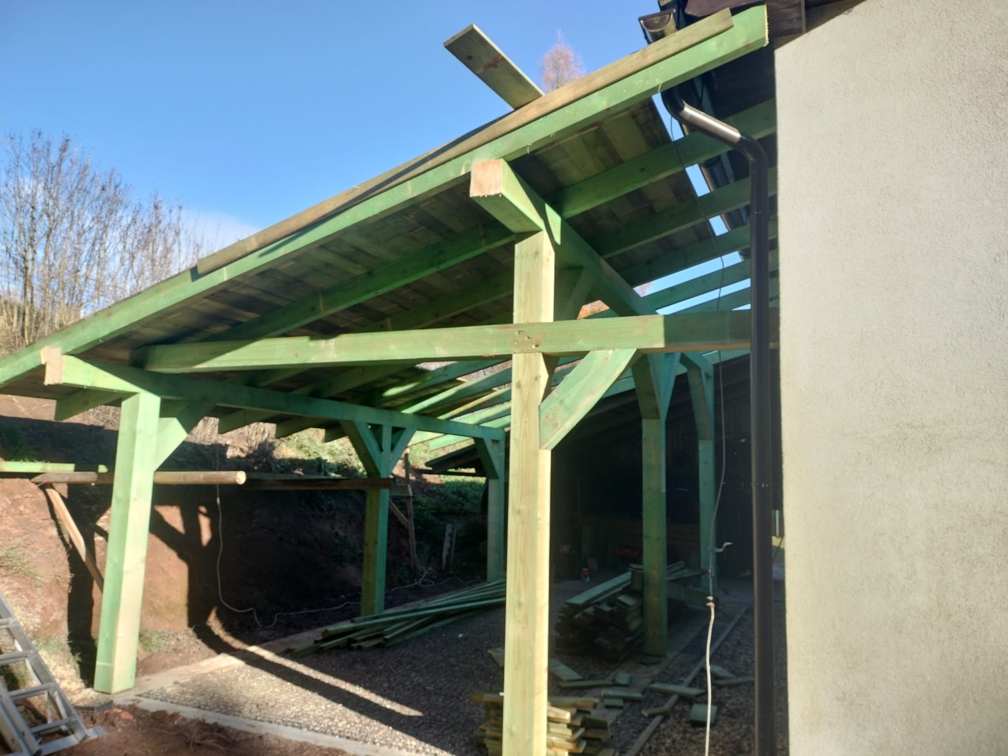 Surowa, zielona konstrukcja drewniana z widoczną więźbą dachową w trakcie budowy, z elementami rynny i fragmentem ściany budynku w tle, w słoneczny dzień.