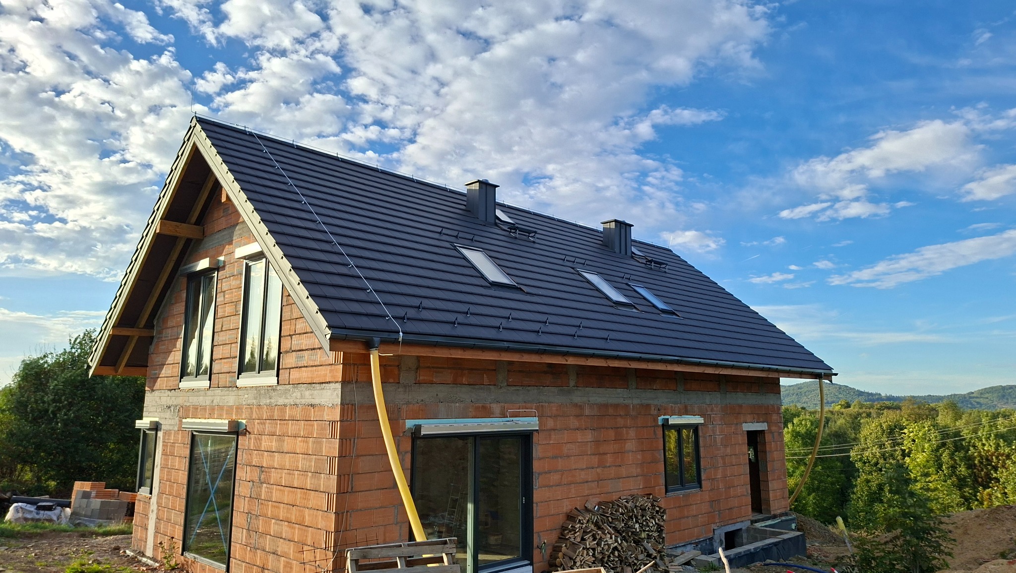 Dom w trakcie budowy z ciemną dachówką, widoczne okna dachowe i kominy. Na pierwszym planie ceglana ściana, w tle błękitne niebo z chmurami i zieleń drzew.