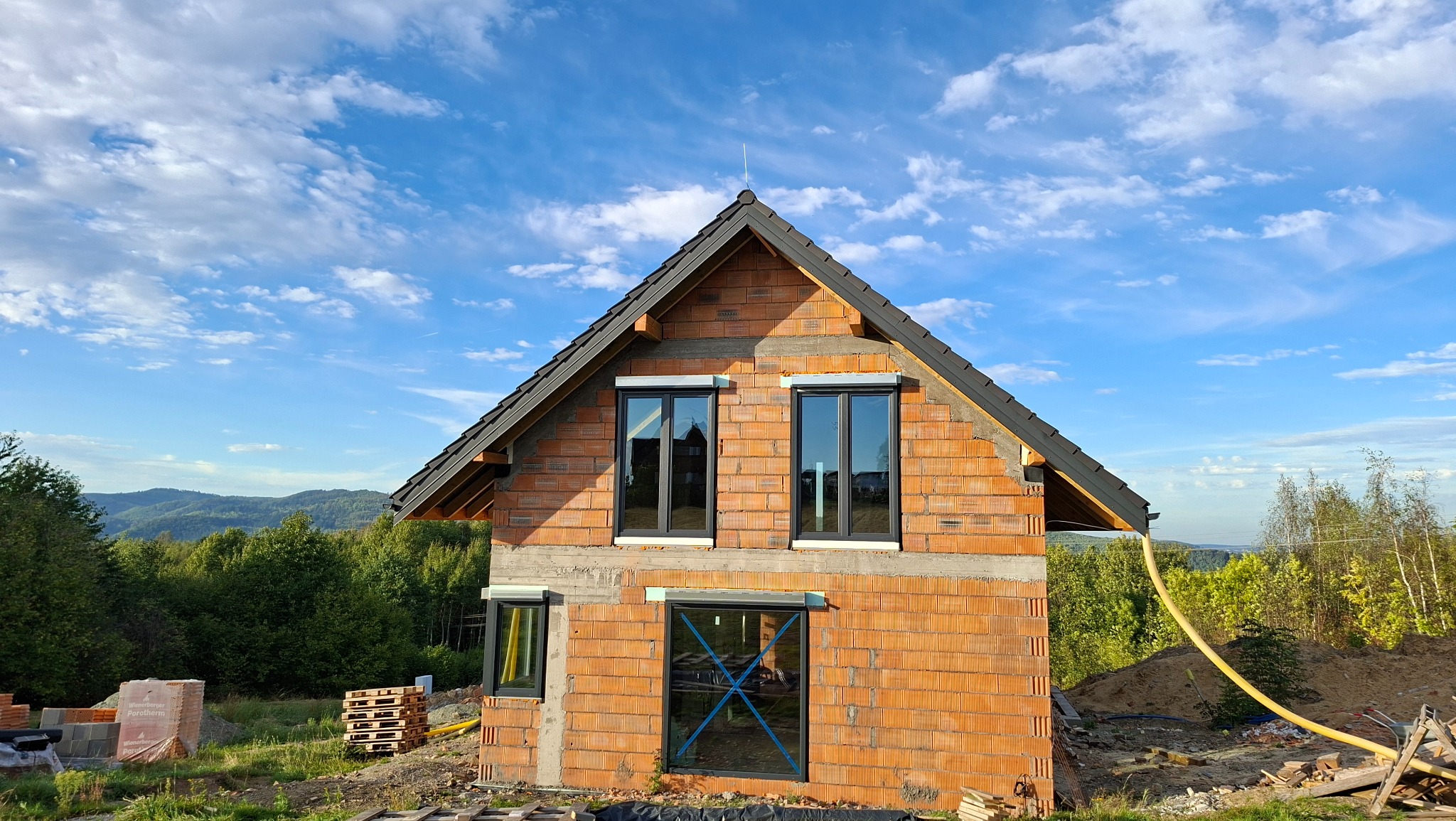 Dom w trakcie budowy z widoczną konstrukcją dachu i oknami. Czerwona cegła, czarne ramy okienne, błękitne niebo w tle. Widok na góry i las.
