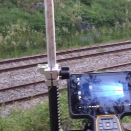 Usługi Geodezyjne GEOAR-2 - Prace geodezyjne przy torach kolejowych: tyczka geodezyjna z niwelatorem i tablet z uruchomionym oprogramowaniem pomiarowym ustawione na tle torowiska i zieleni.