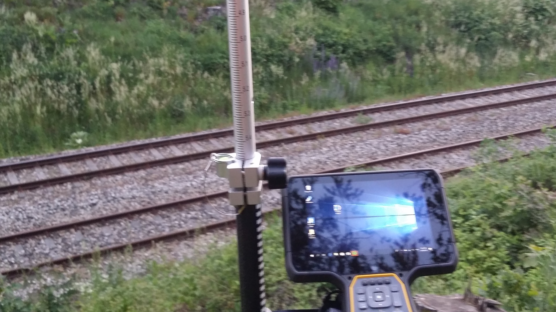 Prace geodezyjne przy torach kolejowych: tyczka geodezyjna z niwelatorem i tablet z uruchomionym oprogramowaniem pomiarowym ustawione na tle torowiska i zieleni.