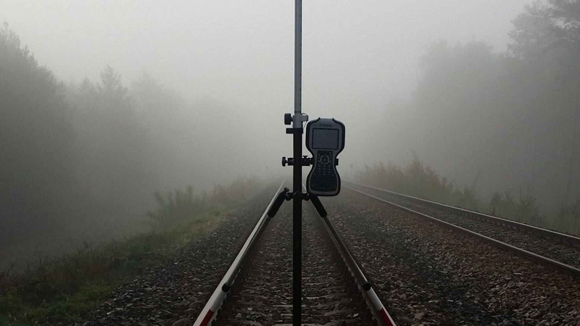 Geodezyjny odbiornik GPS ustawiony na trójnogu, pomiar na torach kolejowych we mgle.