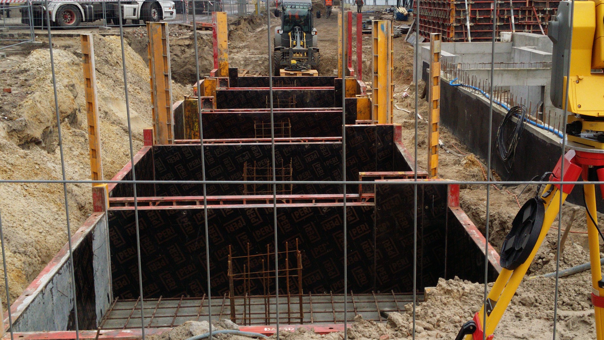 Szeroki wykop pod fundamenty zabezpieczony szalunkiem Peri, widoczna zbrojenie i niwelator na trójnogu, w tle koparka.
