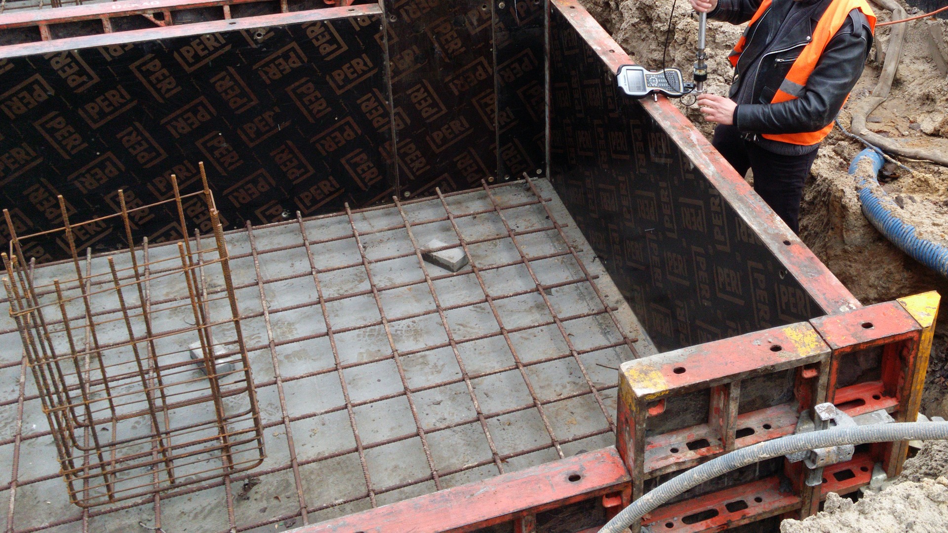 Przygotowany wykop z szalunkiem PERI, zbrojeniem dna i koszem zbrojeniowym, geodeta sprawdza poziom za pomocą niwelatora.