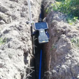 Pomiar geodezyjny wykopu pod instalację rur, widoczny niwelator i fragment niebieskiej rury w wąskim wykopie w ziemi.
