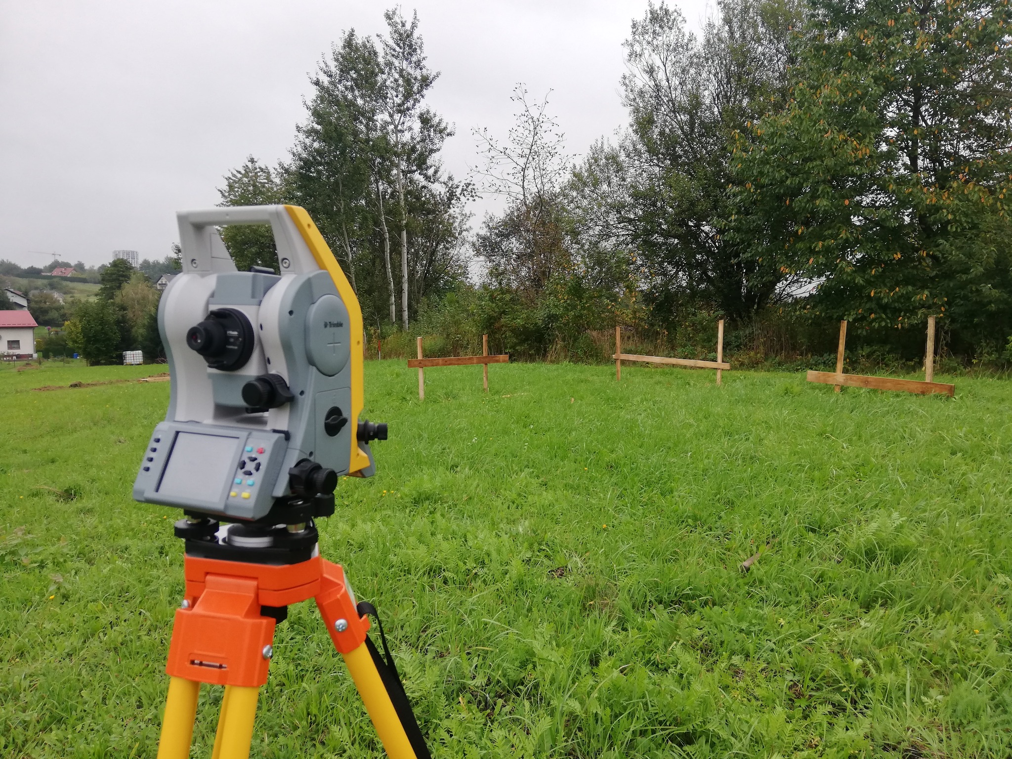 Szary teodolit na pomarańczowo-żółtym statywie, ustawiony na zielonej łące z drewnianymi palikami w tle, pochmurne niebo.