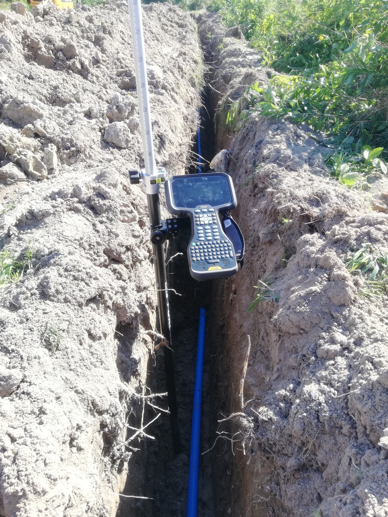 Pomiar geodezyjny wykopu pod instalację rur, widoczny niwelator i fragment niebieskiej rury w wąskim wykopie w ziemi.
