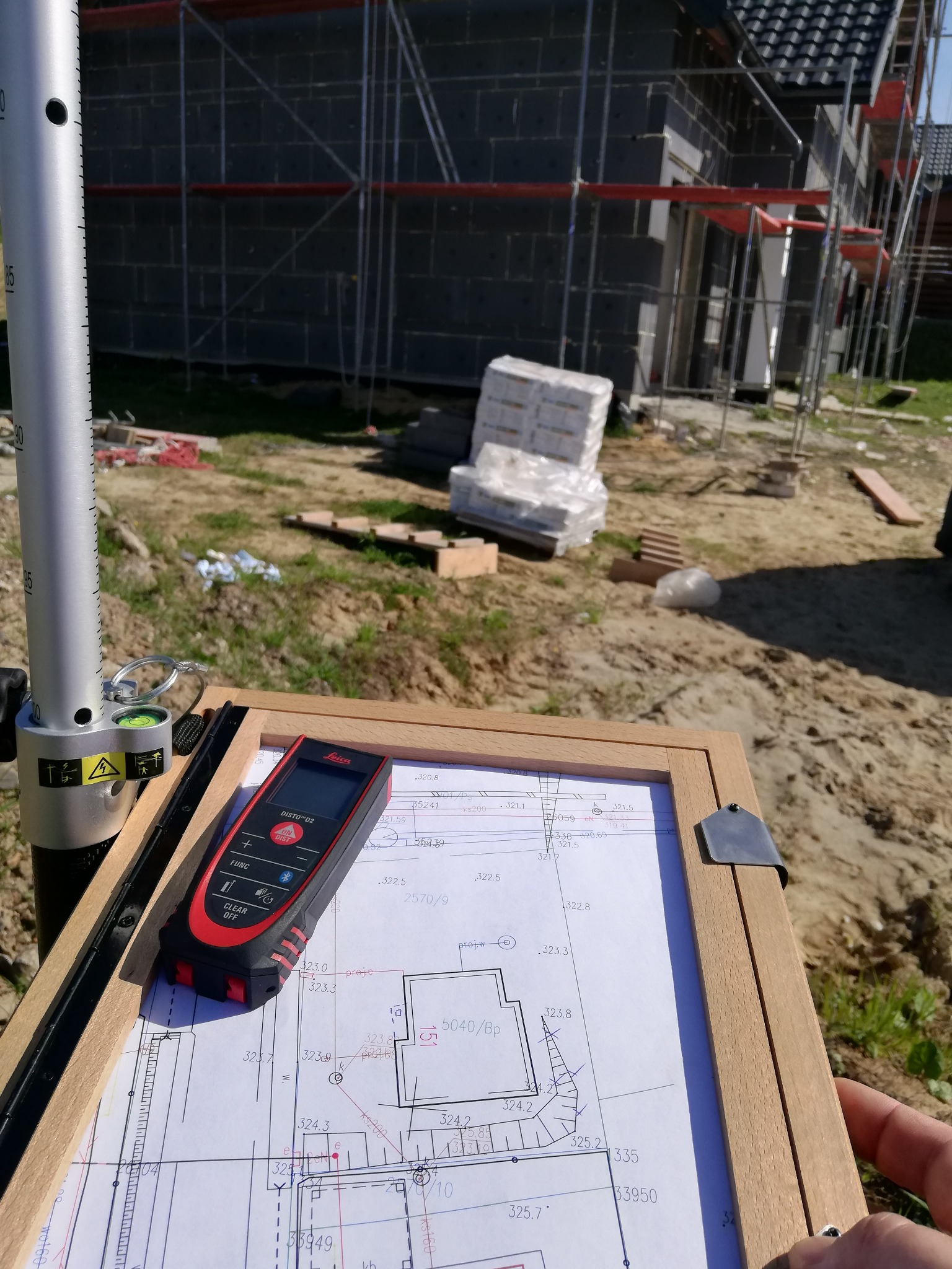 Plan budynku na desce kreślarskiej, dalmierz Leica Disto D2, niwelator w tle budowa domu w trakcie wznoszenia ścian z bloczków.
