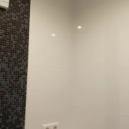 Fragment łazienki z białymi płytkami i mozaiką w odcieniach szarości, widoczne gniazdko elektryczne i półka przy wannie.