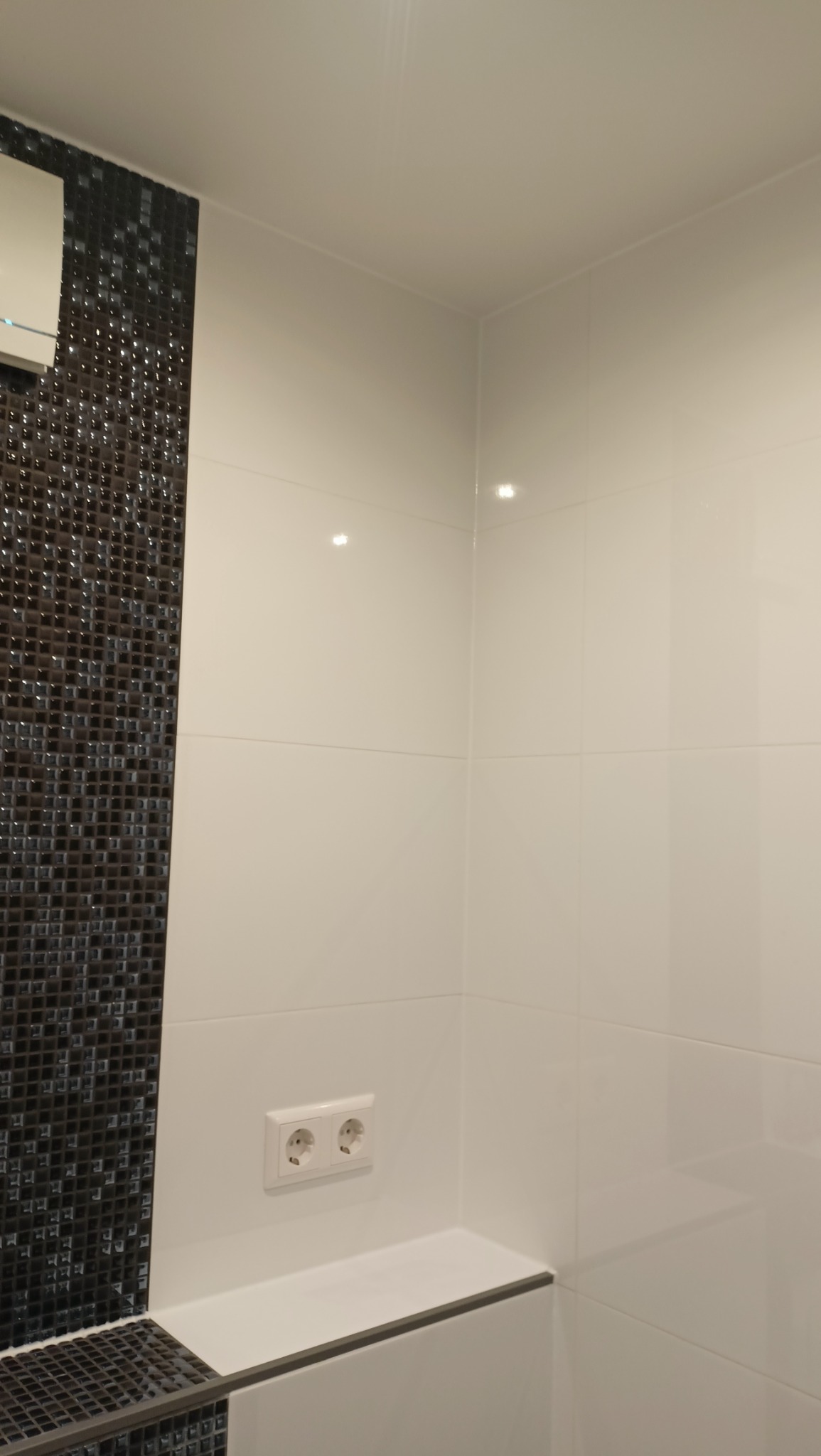 Fragment łazienki z białymi płytkami i mozaiką w odcieniach szarości, widoczne gniazdko elektryczne i półka przy wannie.