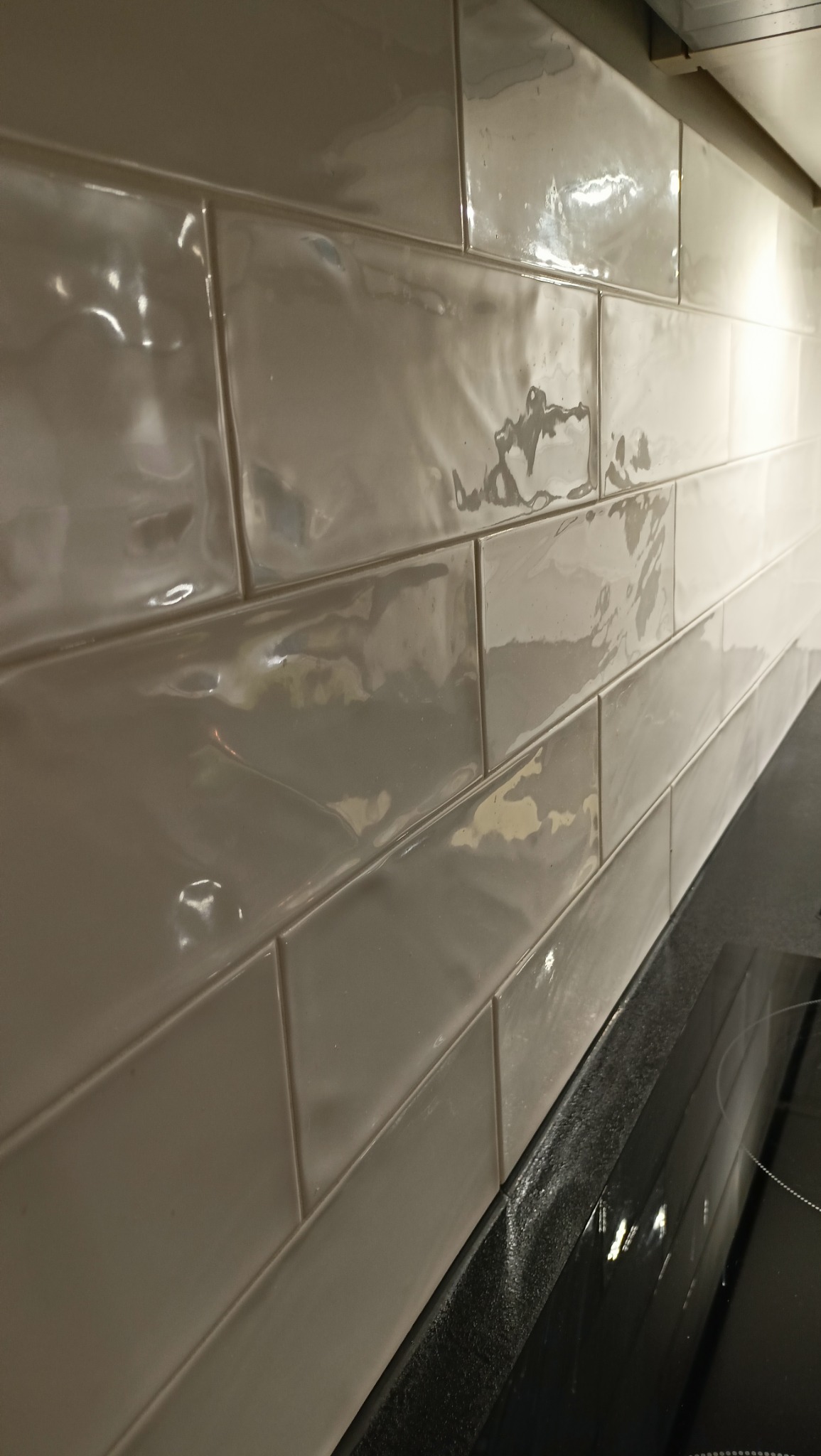 Ściana wyłożona błyszczącymi, kremowymi, prostokątnymi płytkami ceramicznymi, ułożonymi poziomo, z widocznym fragmentem czarnego blatu kuchennego i płyty indukcyjnej.