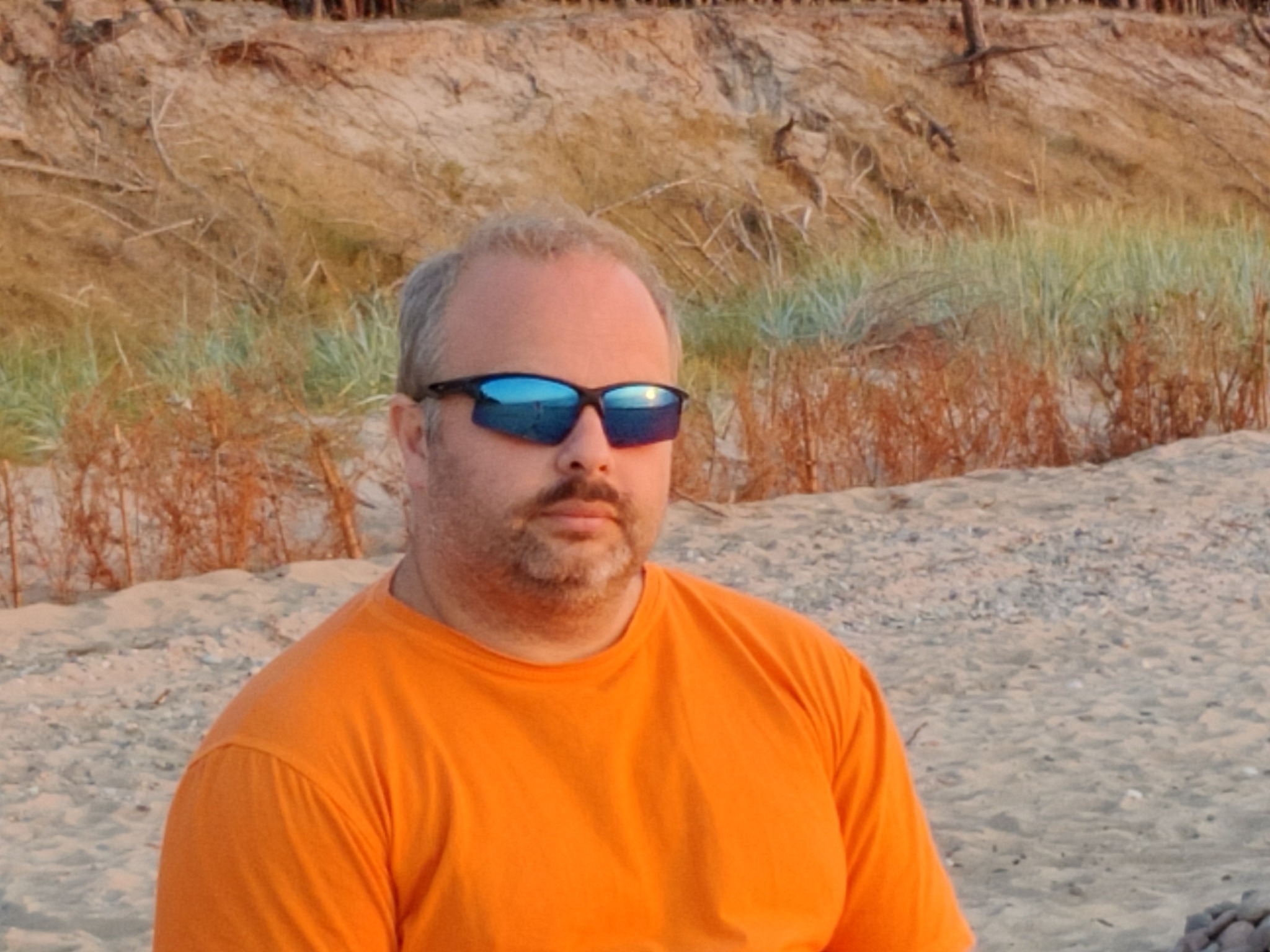 Mężczyzna w pomarańczowej koszulce i okularach przeciwsłonecznych z odbiciem krajobrazu na plaży, stojący na piaszczystym brzegu z wydmami w tle.