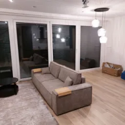 Nowoczesny salon z szarą sofą z drewnianymi elementami, widok na kuchnię przez przesuwne drzwi balkonowe, drewniana podłoga i dywan z abstrakcyjnym wzorem, wiszące lampy kuliste.