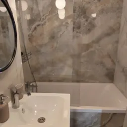 Nowoczesna łazienka z marmurowymi płytkami, wiszącą lampą kulową, białą umywalką z szafką i wanną za szklanym parawanem.