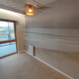 Pokój z drewnianą podłogą ułożoną w jodełkę, ściana z malowidłem imitującym plażę, nowoczesna lampa wisząca z ażurowym kloszem i balkon z widokiem na plac budowy.