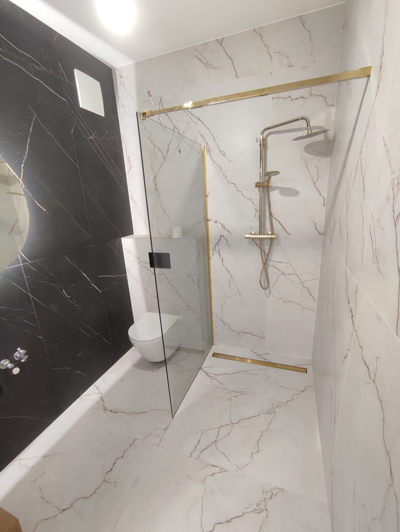 Nowoczesna łazienka z kabiną prysznicową typu walk-in, wykończona płytkami imitującymi marmur w odcieniach bieli i szarości, z elementami w kolorze złotym, takimi jak rama kabiny i odpływ liniowy...