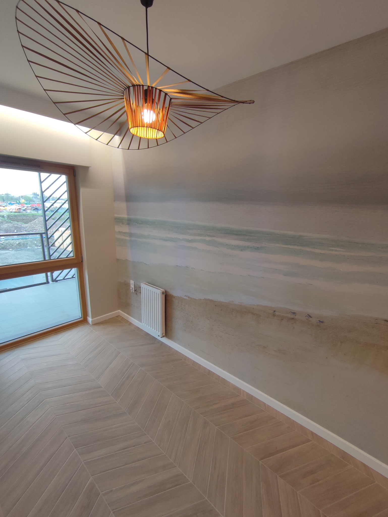 Pokój z drewnianą podłogą ułożoną w jodełkę, ściana z malowidłem imitującym plażę, nowoczesna lampa wisząca z ażurowym kloszem i balkon z widokiem na plac budowy.