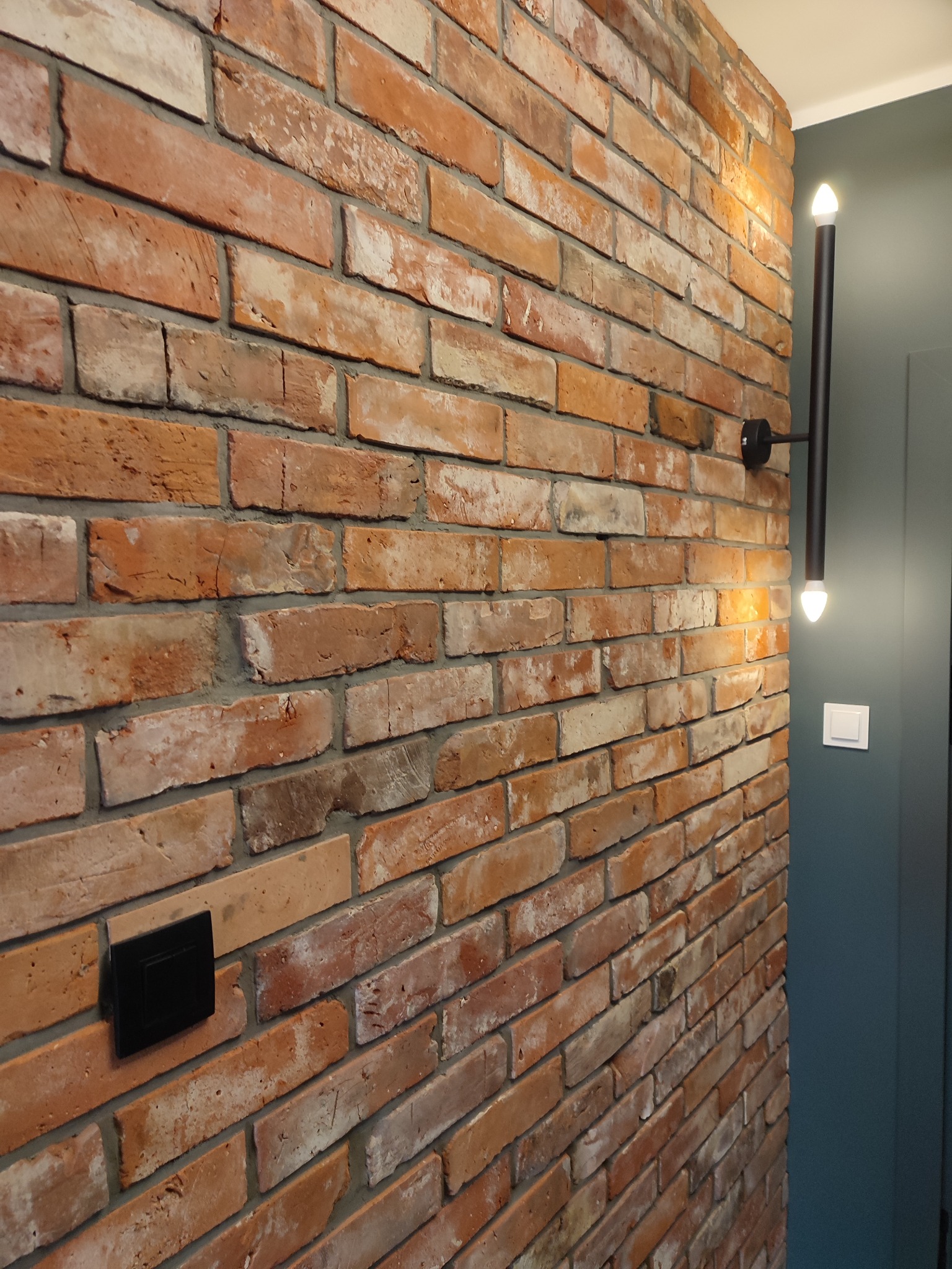 Ściana z cegły klinkierowej w odcieniach czerwieni i bieli, oświetlona nowoczesną, czarną lampą ścienną z dwoma punktami świetlnymi, z widocznym czarnym gniazdkiem elektrycznym i białym włącznikiem...