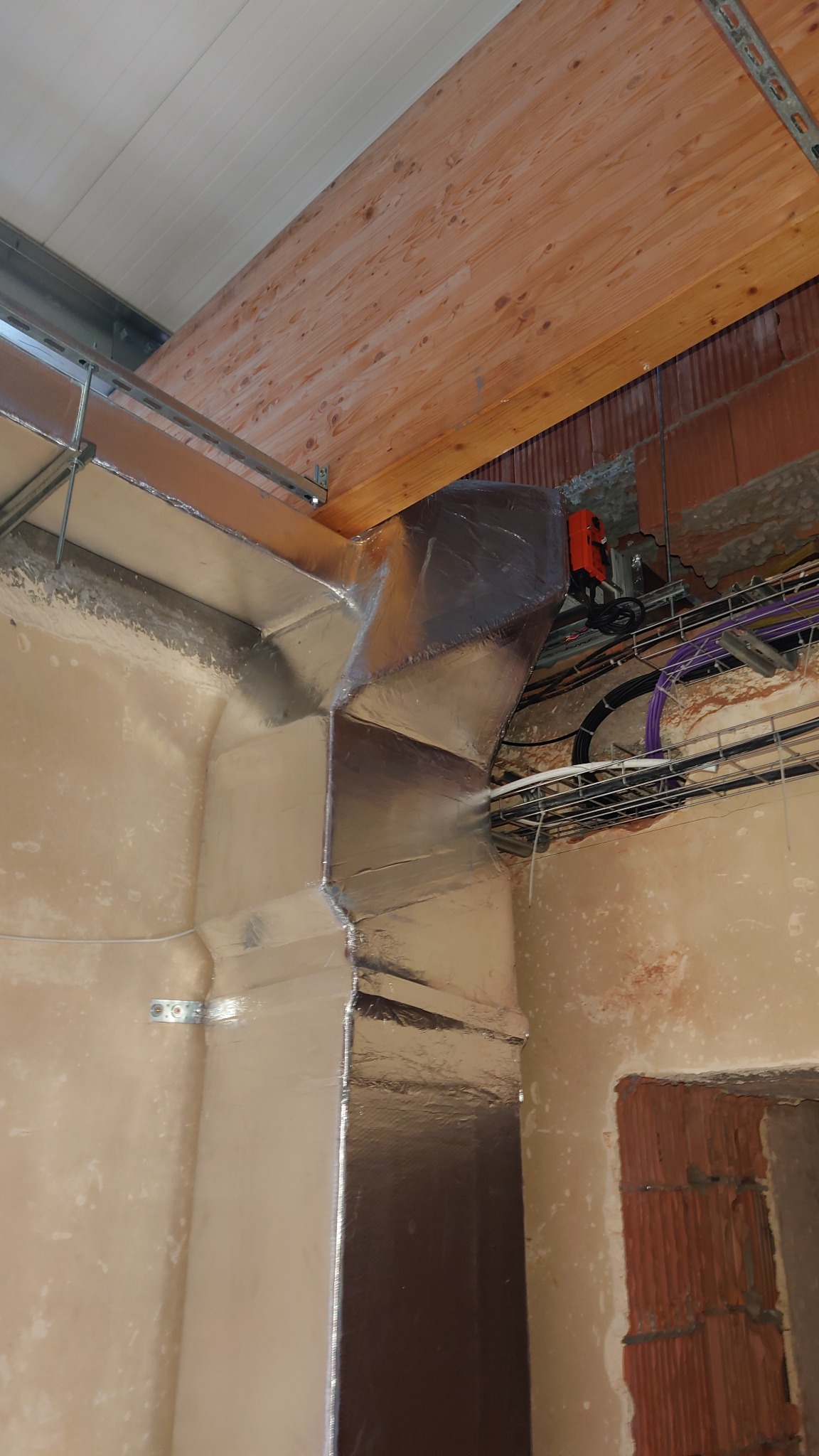 Kanciasty kanał wentylacyjny w izolacji aluminiowej, zamontowany w rogu pomieszczenia w trakcie wykańczania, widoczne przewody i elementy konstrukcyjne budynku.