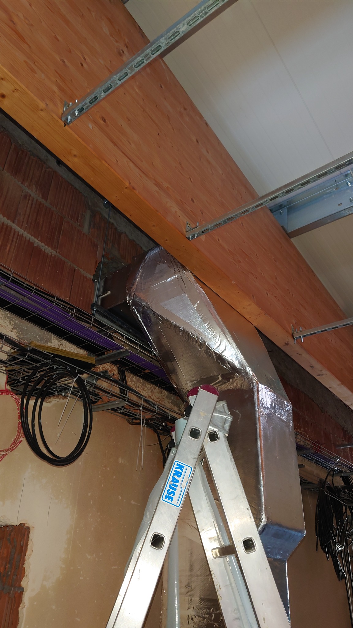 Montaż izolowanego kanału wentylacyjnego na drabine Krause w surowym wnętrzu z widocznymi kablami i elementami konstrukcyjnymi.