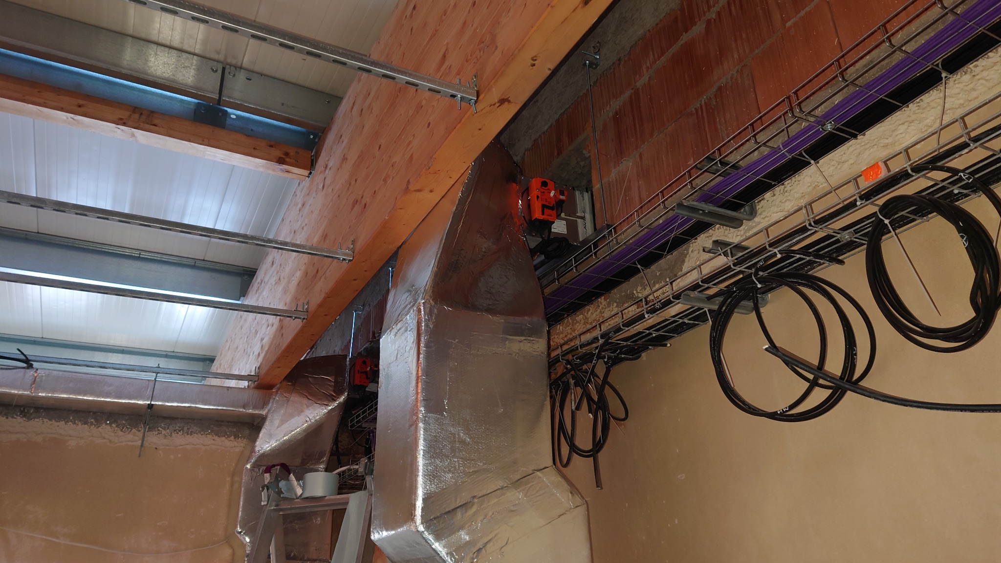 Szczegółowe ujęcie instalacji kanałów wentylacyjnych owiniętych folią aluminiową, z widocznymi elementami mocującymi do drewnianej konstrukcji stropu oraz kablami w korytkach kablowych pod sufitem.