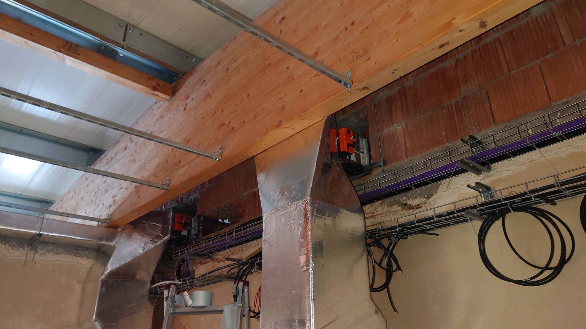 Metalowe kanały wentylacyjne z izolacją, zamontowane pod drewnianą konstrukcją stropu, widoczne kable i instalacje elektryczne na drabinkach kablowych.